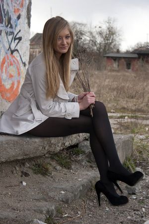Leggy and sexy blonde posing outdoors near some abandoned building in black pantyhose, heels and coat.