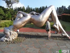 Funny fantastical giant silver colored statue of a striking nude with man posing for interesting picture between her sexy legs pretending
